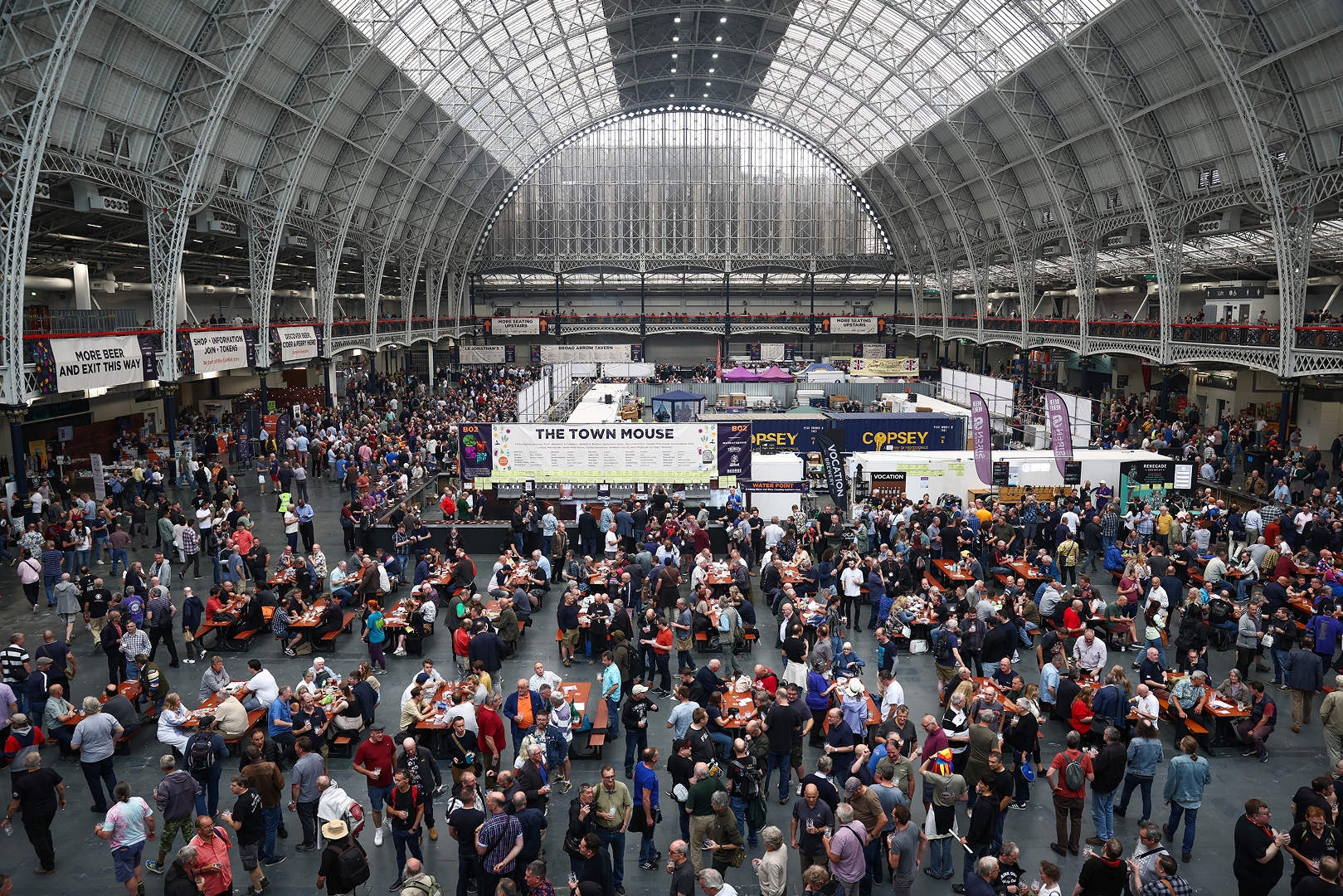 A wide, high-angle view of a bustling beer festival inside a large, glass-roofed hall with hundreds of people sitting at communal tables and visiting various brewery stands like 'The Town Mouse'.