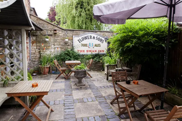 A pub garden with tables and a sign on the garden wall reading: FLOWER & SONS - Fine ALES - THE SWAN INN
